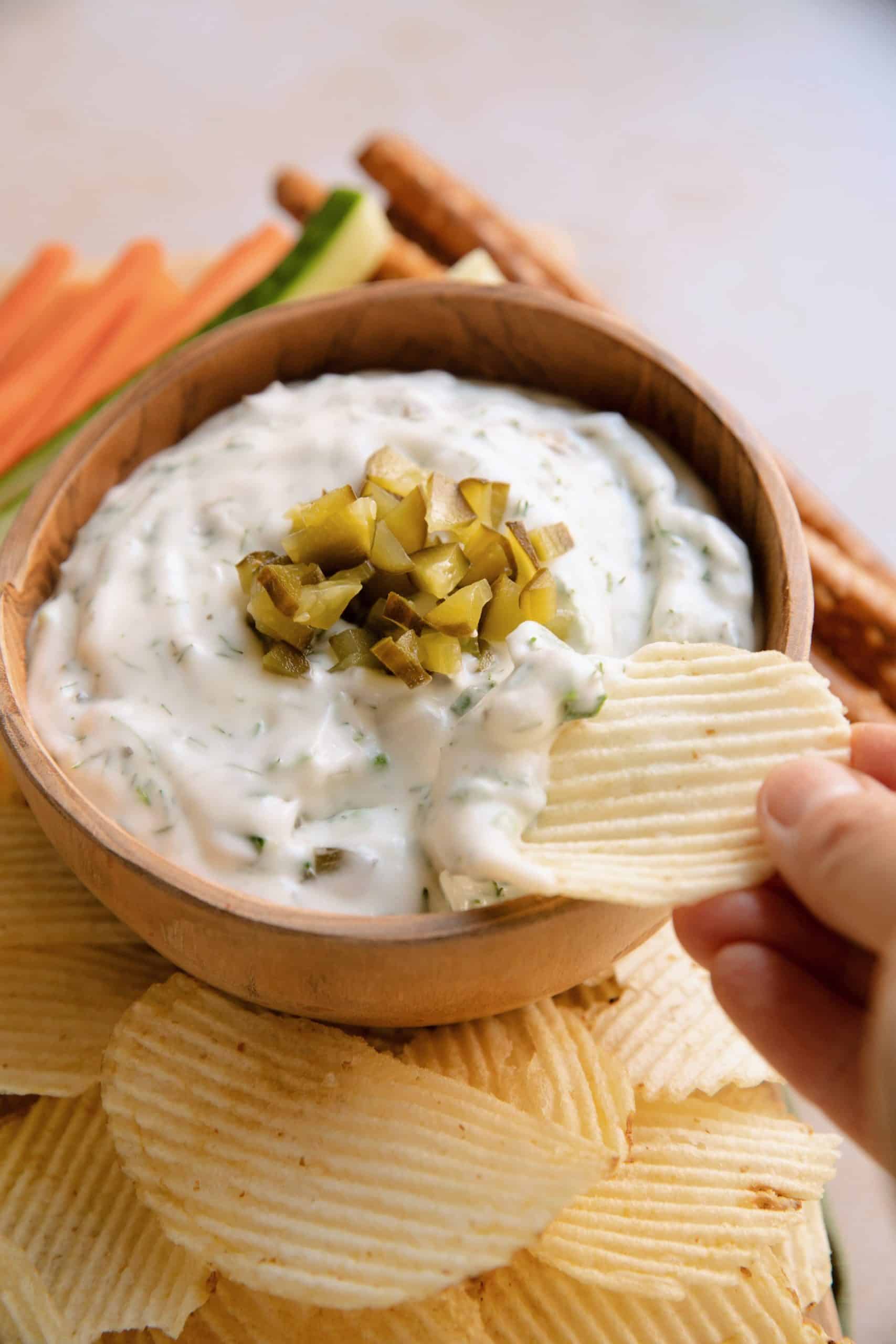 a hand dipping a ruffled chip into a bowl of dill pickle dip